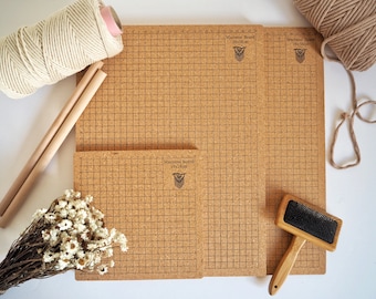 May include: Three cork macrame boards with grid lines and the text "Macrame Board" and measurements in inches. The boards are on a white surface with a ball of yarn, two wooden dowels, a bunch of dried flowers, and a wooden comb.