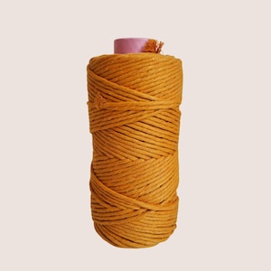 Könnte beinhalten: Eine Spule Senfgelbes Makramee-Garn. Das Garn ist eng um einen zylindrischen Kern gewickelt, der mit einer rosa Kappe und einer kleinen, orangefarbenen Quaste versehen ist. Das Garn hat ein strukturiertes, gewebtes Aussehen, geeignet für Bastelprojekte.