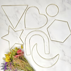 May include: A set of six gold metal hoops in various shapes, including a heart, a circle, a star, a triangle, a crescent moon, and a hexagon. The hoops are arranged on a white surface.