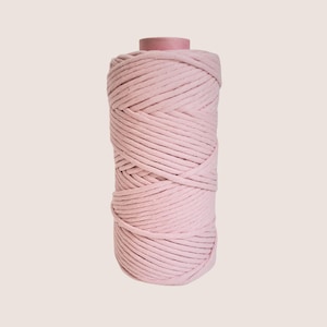 Könnte beinhalten: Eine Spule hellrosa Makramee-Kordel. Die Kordel ist eng um einen Pappkern gewickelt und bildet eine zylindrische Form. Die Farbe ist ein weiches, gedämpftes Rosa, das für verschiedene Bastelprojekte geeignet ist. Die Kordel scheint aus einem weichen, strapazierfähigen Material zu bestehen.