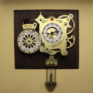 Op de afbeelding: Een goudkleurige klok met ingewikkelde tandwielen en een slinger. De wijzerplaat van de klok heeft Romeinse cijfers en de woorden "HOURS" en "MINUTES" staan op de wijzerplaat gedrukt.