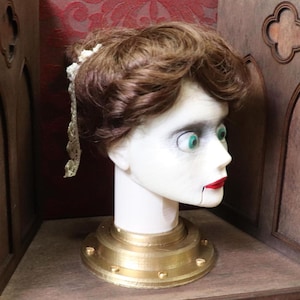 May include: A vintage-style doll head with brown hair, green eyes, and red lips. The head is mounted on a white neck and a gold-colored base. A lace hair accessory is attached to the hair. The doll head is displayed in a wooden cabinet.
