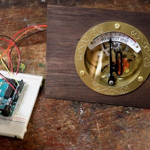 May include: A blue Arduino microcontroller board connected to a breadboard with wires. The board is connected to a wooden base with a brass clock face that reads "YOUR TEXT HERE".