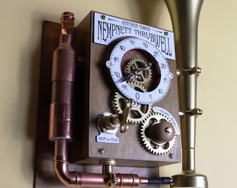Puede incluir: Un temporizador de cocina de estilo steampunk con una campana de latón, tuberías de cobre y una esfera de reloj con engranajes. El temporizador está etiquetado como "Kitchen Timer Nempnett Thruibwell".