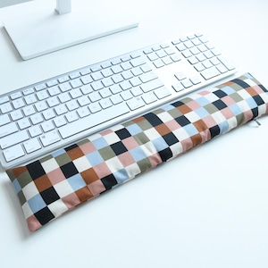 Puede incluir: Un reposamuñecas rectangular para teclado con un patrón de cuadros multicolor en tonos negro, blanco, azul claro, rosa empolvado y verde oliva. La funda de tela tiene una pequeña cremallera en el lado derecho y se sitúa bajo el teclado.