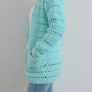 May include: A light blue crocheted cardigan with long sleeves and pockets. The cardigan is made with a delicate open stitch pattern.