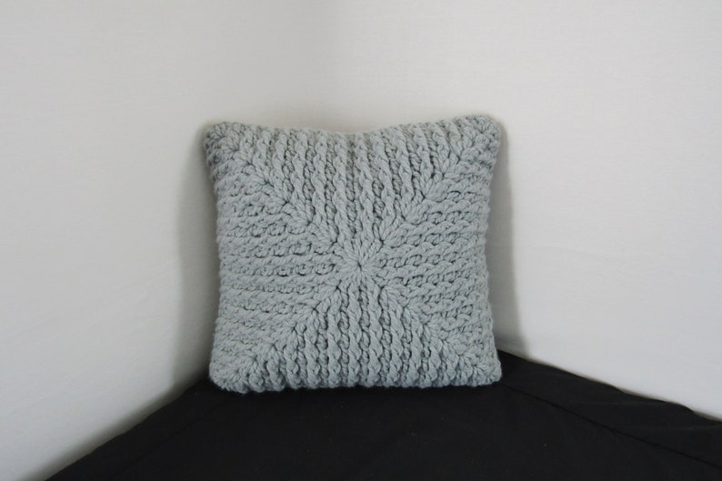 K&ouml;nnte beinhalten: Ein graues gestricktes Kissen mit einem strukturierten, gewebten Design. Das Kissen ist quadratisch und hat ein weiches, flauschiges Aussehen.