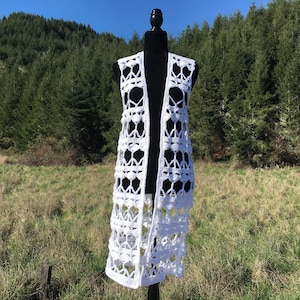 May include: A white, open-front crochet vest with a geometric pattern. The long vest is displayed on a black mannequin against a backdrop of a grassy field and a forest of evergreen trees. The vest's design features openwork squares and textured details.