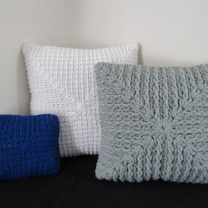 K&ouml;nnte beinhalten: Drei gestrickte Kissen, eines wei&szlig;, eines grau und eines blau. Die Kissen sind alle quadratisch und haben ein strukturiertes Muster.