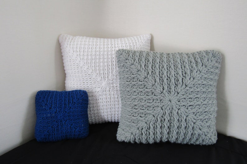 K&ouml;nnte beinhalten: Drei gestrickte Kissen, eines wei&szlig;, eines grau und eines blau. Die Kissen sind alle quadratisch und haben ein strukturiertes Muster.
