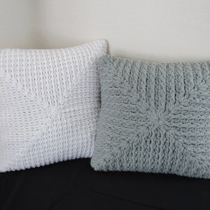 K&ouml;nnte beinhalten: Zwei gestrickte Kissen, eines wei&szlig; und eines grau, mit einem strukturierten Sternmuster. Die Kissen liegen auf einer schwarzen Oberfl&auml;che.