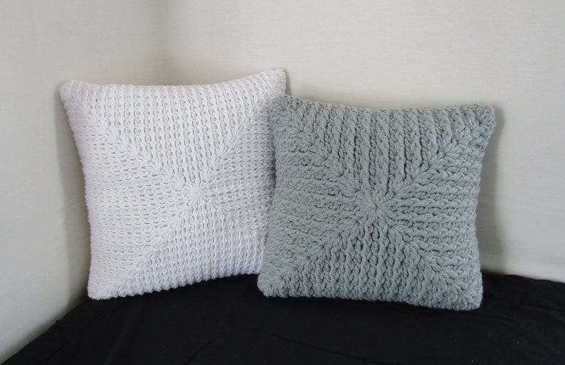 K&ouml;nnte beinhalten: Zwei gestrickte Kissen, eines wei&szlig; und eines grau, mit einem strukturierten Sternmuster. Die Kissen liegen auf einer schwarzen Oberfl&auml;che.