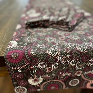 May include: A dark brown table runner with a floral pattern featuring pink, white, and gray flowers. The runner has a scalloped edge and is made of a soft, cotton fabric.