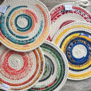 Peut inclure: Ensemble de dessous-de-plat circulaires faits à la main avec un motif en spirale. Chaque dessous-de-plat présente une bordure en corde naturelle et un centre en tissu coloré, avec des motifs rouges, bleus, verts et jaunes. Ils ont une boucle pour suspendre.