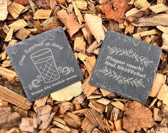 May include: Two slate coasters with engraved sayings. The first coaster says "One apple a day keeps the doctor away" with a glass of cider. The second coaster says "Finger weg! Mei Stoffche!"