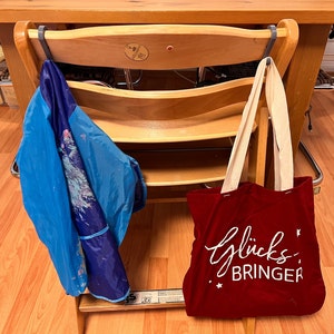 Könnte beinhalten: Eine rote Tragetasche mit dem Schriftzug "Glücksbringer" in weißen Buchstaben hängt an einem Holzstuhl. Ein blaues und weiß gemustertes Tuch hängt ebenfalls an dem Stuhl.