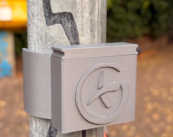 Puede incluir: Una caja rectangular gris con un emblema circular y un diseño estilizado está sujeta a un poste metálico. La caja está asegurada con una banda gris a juego. El fondo muestra una vista borrosa de árboles y follaje.