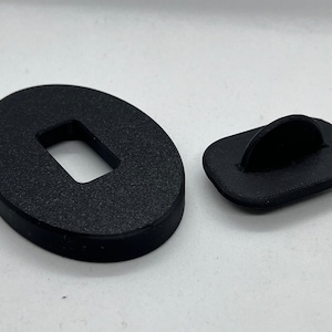 May include: Two black plastic pieces, one oval-shaped with a rectangular hole in the center, and the other rectangular with a curved hook on top.