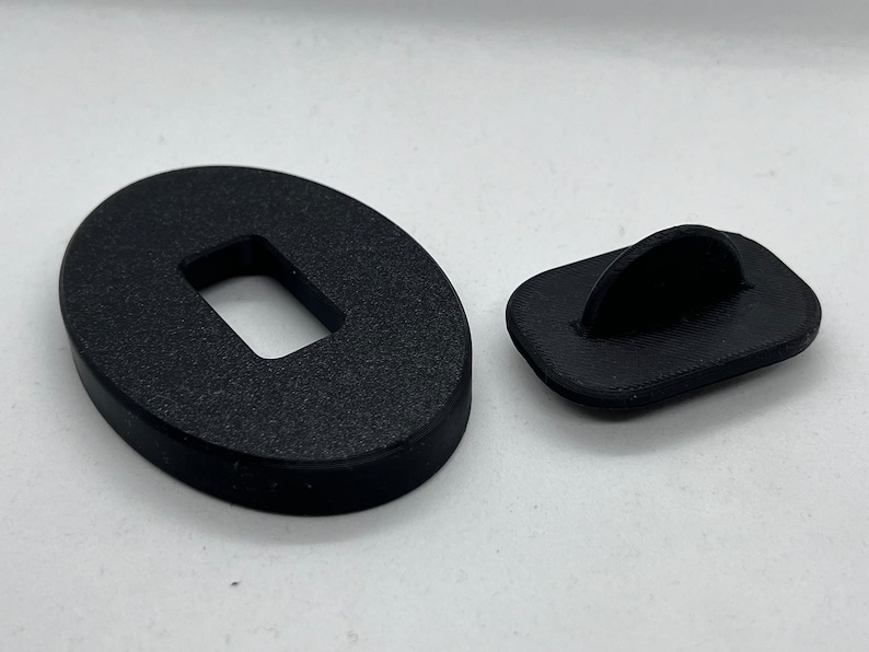May include: Two black plastic pieces, one oval-shaped with a rectangular hole in the center, and the other rectangular with a curved hook on top.