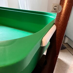 K&ouml;nnte beinhalten: Ein gr&uuml;ner Plastik-Aufbewahrungskorb mit einem wei&szlig;en Griff, der an einem braunen Holzrahmen befestigt ist.
