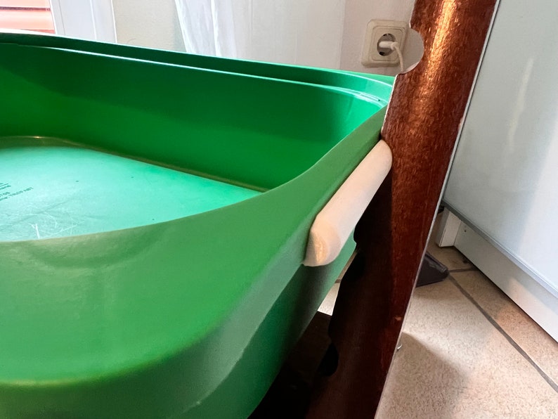 K&ouml;nnte beinhalten: Ein gr&uuml;ner Plastik-Aufbewahrungskorb mit einem wei&szlig;en Griff, der an einem braunen Holzrahmen befestigt ist.