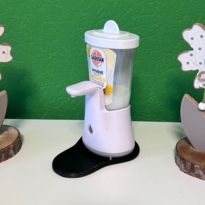 May include: White automatic soap dispenser with a black base. The dispenser has a clear plastic bottle with a label that reads "Sagrotan Hygiene Seife Ultra Care".