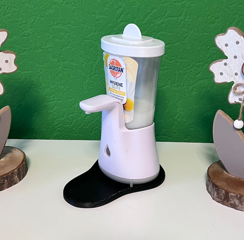 May include: White automatic soap dispenser with a black base. The dispenser has a clear plastic bottle with a label that reads "Sagrotan Hygiene Seife Ultra Care".