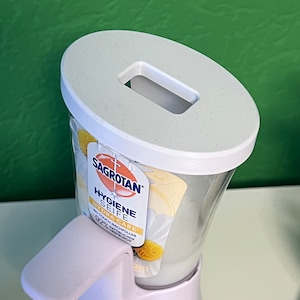 May include: White plastic soap dispenser with a white pump and a clear bottle. The bottle is labeled "Sagrotan Hygiene Seife Hydra Care" with a daisy design. The dispenser is on a white base.