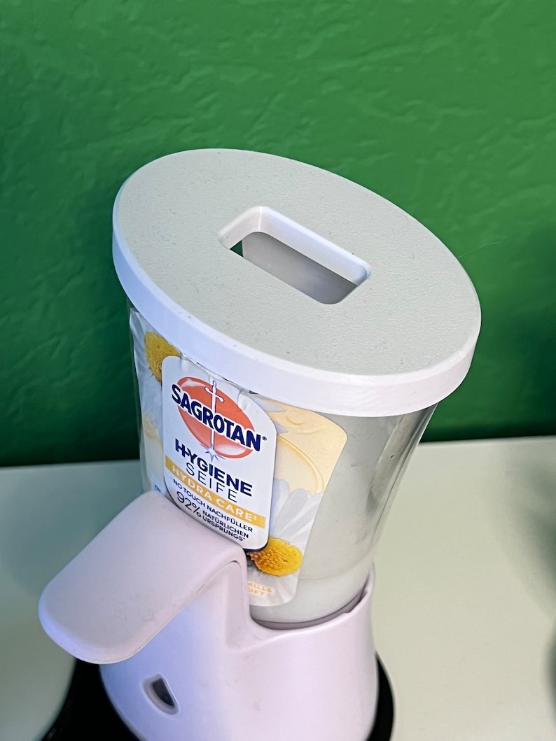 May include: White plastic soap dispenser with a white pump and a clear bottle. The bottle is labeled "Sagrotan Hygiene Seife Hydra Care" with a daisy design. The dispenser is on a white base.