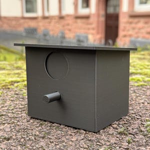 May include: A dark gray birdhouse with a circular entrance hole and a small perch. The birdhouse is box-shaped with a flat roof and is sitting on a textured surface. The background features a building with arched windows.