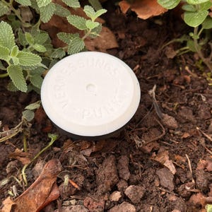 May include: A white, circular garden sensor with the text "VERMESSUNGSPUNKT" on top. The sensor is set in dark soil, surrounded by green plants and brown mulch.