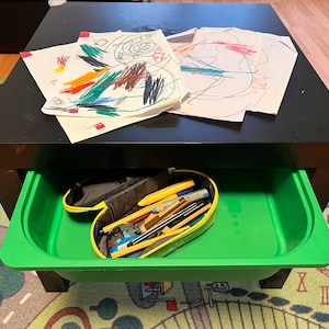 May include: A black table with a green drawer. The drawer is open and contains a yellow pencil case with pencils and pens inside. There are three pieces of paper with colorful drawings on the table.