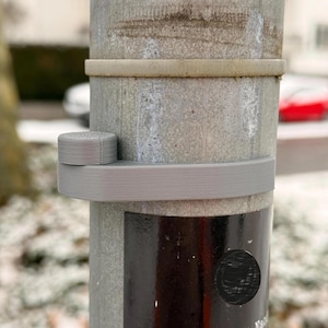May include: A gray, 3D-printed bracket and cap are attached to a weathered metal pole. The bracket wraps around the pole, and the cap sits on top. The pole has a dark, glossy section with a circular detail.