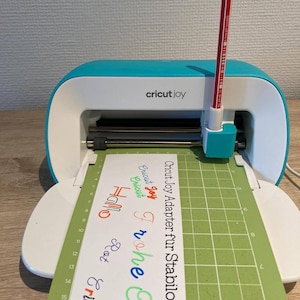 May include: A turquoise Cricut Joy cutting machine with a green cutting mat and a red pen in the holder. The cutting mat has a grid pattern and the text "Cricut Joy Adapter fur Stabilos" is printed on the mat. The text "Cricut Joy" is printed on the machine.