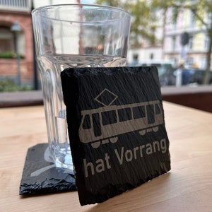 May include: A black slate coaster featuring a white graphic of a tram and the text "hat Vorrang". The coaster is placed on a wooden table beside a glass of water.