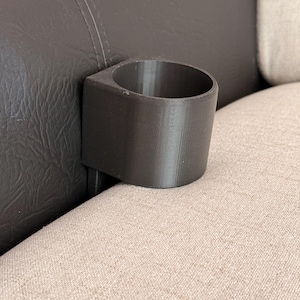Puede incluir: Un portavasos negro, impreso en 3D, fijado al lado de un sofá. El portavasos es cilíndrico con una base cuadrada y está posicionado contra un cojín de cuero marrón oscuro y un cojín de tela beige.