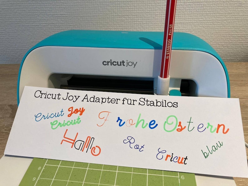 May include: A Cricut Joy cutting machine with a red Stabilo pen inserted. The machine is on a green cutting mat with a white card that says "Cricut Joy Adapter fur Stabilos" and has several words written on it in different colors, including "Cricut Joy", "Frohe Ostern", "Hallo", "Rot", "Cricut", and "blau".