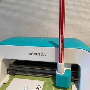 May include: A red pen with white stripes is inserted into the pen holder of a Cricut Joy cutting machine. The machine is turquoise blue and white. The machine is on a green cutting mat with a white card on top of it. The card has the words "Cricut Joy" written on it in green, orange, and pink.