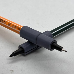 May include: Two gray pen grips with black tips, one on top of the other, with a yellow pencil with black text that reads "NO. 88/A1 pp" in the background.