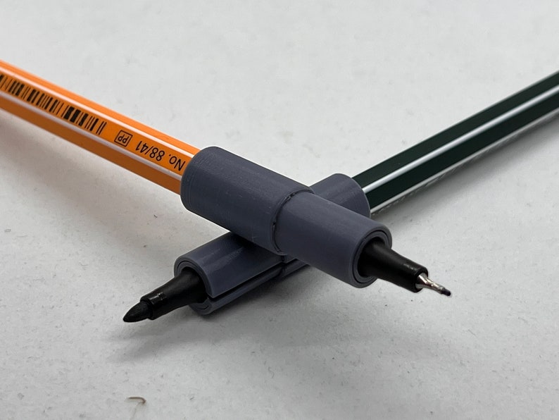 May include: Two gray pen grips with black tips, one on top of the other, with a yellow pencil with black text that reads "NO. 88/A1 pp" in the background.