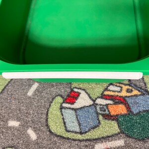 K&ouml;nnte beinhalten: Ein gr&uuml;ner Plastik-Aufbewahrungsbeh&auml;lter mit einem wei&szlig;en Klebestreifen am oberen Rand. Der Beh&auml;lter steht auf einem bunten Teppich.