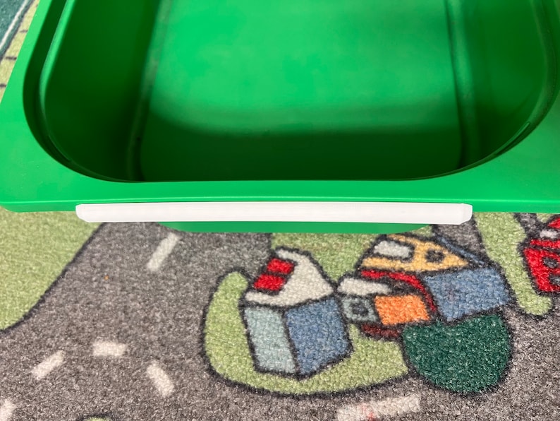 K&ouml;nnte beinhalten: Ein gr&uuml;ner Plastik-Aufbewahrungsbeh&auml;lter mit einem wei&szlig;en Klebestreifen am oberen Rand. Der Beh&auml;lter steht auf einem bunten Teppich.