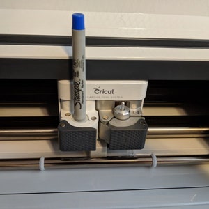 Könnte beinhalten: Eine weiße Cricut-Schneidemaschine mit einem blauen Sharpie-Stift, der in den Werkzeughalter eingesetzt ist. Die Maschine hat einen silbernen Metallwagen und ein schwarz-graues, strukturiertes Gehäuse.