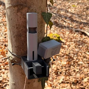 May include: A gray, 3D-printed device attached to a tree trunk. The device has a cylindrical component with a rectangular cutout, a rectangular box, and a smaller square component. The device is mounted with black zip ties and a pink wire.