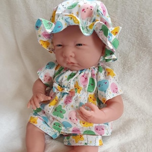 May include: A doll wearing a white sun hat and matching short-sleeved shirt and shorts set. The clothing features a colourful pattern of cartoon balloons and stars. The doll is posed on a white surface.