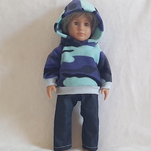 May include: A doll dressed in a blue, purple, and teal camouflage hooded sweatshirt and dark blue denim trousers. The sweatshirt has a white hem and a teal-lined hood. The doll stands on a white surface.
