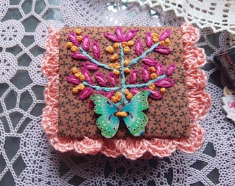 May include: A small, square, brown fabric box with a floral embroidery design in pink, yellow, and blue. A wooden butterfly is attached to the front. The box is surrounded by a pink, crocheted border.