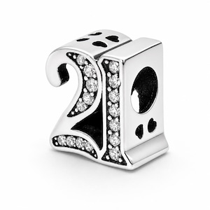 Op de afbeelding: Een zilverkleurige bedel in de vorm van het getal 21, voorzien van heldere, fonkelende steentjes ingelegd aan de voorzijde van beide cijfers. De boven- en zijkanten zijn versierd met kleine, uitgesneden hartvormen en een rond gat is zichtbaar.