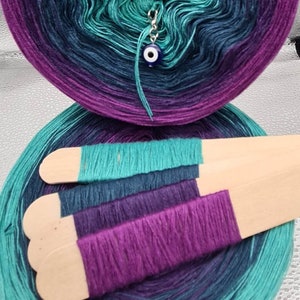 May include: A skein of yarn in shades of teal, purple, and navy blue. The yarn is wound into a ball and has a small blue evil eye charm attached to it. The yarn is displayed on a wooden dowel.
