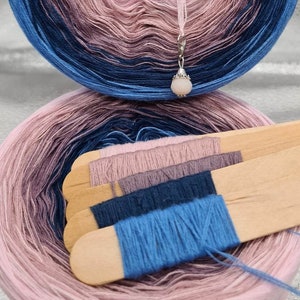 Könnte beinhalten: Eine Garnrolle mit Ombre-Farben in den Farben Rosa, Lila, Blau und Marine. Das Garn ist zu einer Kugel gewickelt und hat einen kleinen silbernen Anhänger. Das Garn wird auf einem Holzstab mit Garnproben in verschiedenen Farben gezeigt.
